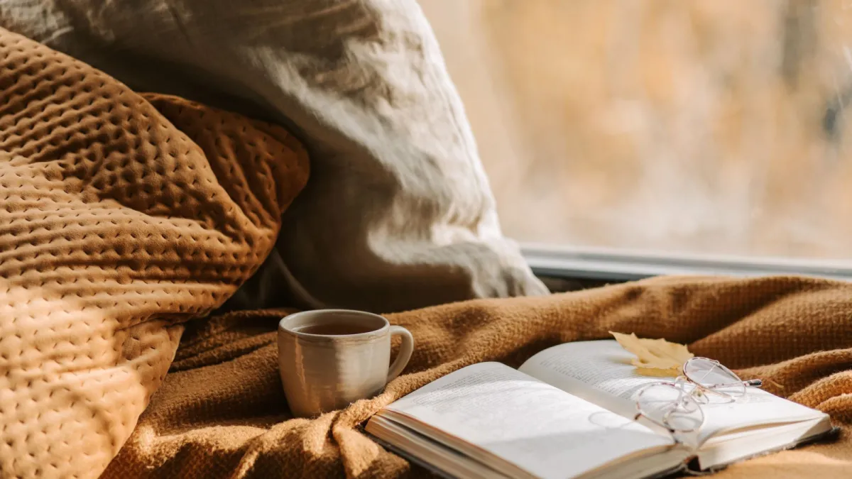 Pencere kenarında örgü battaniyenin üzerinde bir fincan çay ve gözlüklü açık bir kitap.