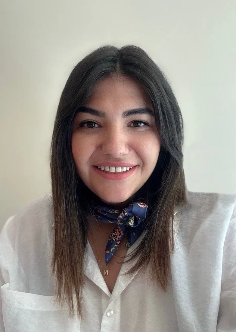 Klinik Psikolog Nilay Burhanoğlu; beyaz gömlek ve lacivert çiçekli fular takmış, açık renk bir duvarın önünde gülümsüyor.